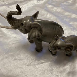 May include: A glossy gray ceramic figurine set featuring a large elephant with a raised trunk and long white tusks, accompanied by a smaller elephant walking beside it. Both animals have detailed ears, textured skin, and a smooth, reflective finish.