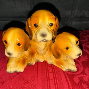May include: A ceramic figurine featuring three golden-brown puppies with dark eyes and floppy ears. The central puppy sits upright while the two puppies on either side are positioned in a reclining pose, all connected as a single decorative piece.