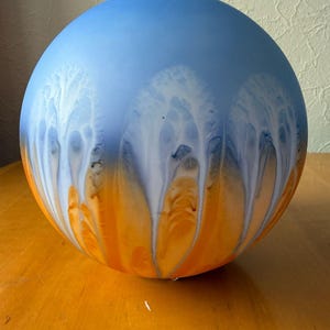 May include: A spherical glass object featuring a smooth gradient transition from a light blue top to a vibrant orange base. The surface displays a white, tree-like, or fern-like pattern that cascades vertically from the blue section into the orange.