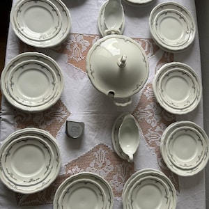 May include: A collection of off-white ceramic dinnerware featuring a delicate gray floral border pattern. The set includes a large lidded tureen, two gravy boats, and ten plates of varying sizes. A small silver measuring tape is placed on the table.