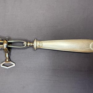 May include: A silver-toned metal tool featuring a long, smooth handle with a shell-like motif at the end. The opposite end consists of a mechanical clamp mechanism with three prongs and a diamond-shaped adjustment screw for tightening the grip.