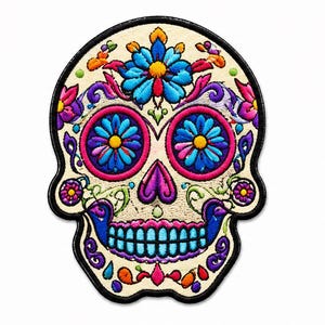 Könnte beinhalten: Ein gestickter Aufnäher in Totenkopfform mit cremefarbener Basis. Das Design zeigt leuchtend blaue, rosa, violette und orangefarbene Blumenmuster um die Augenhöhlen und auf der Stirn, mit einem schwarzen Rand, der die gesamte Totenkopfform umreißt.