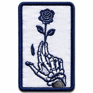 May include: A rectangular embroidered patch featuring a white background with a dark blue border. The design depicts a skeletal hand holding a single dark blue rose on a stem, with a small dark blue petal falling to the left of the hand.