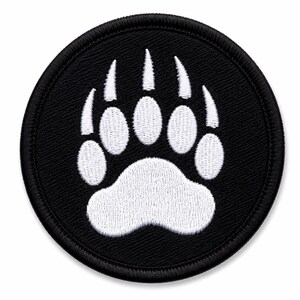 Op de afbeelding: Een ronde geborduurde patch met een zwarte achtergrond en een witte berenpootafdruk in het midden. De pootafdruk bevat vijf duidelijke klauwsporen en een grote centrale voetzool, allemaal uitgevoerd in wit garen met een gestikte rand.