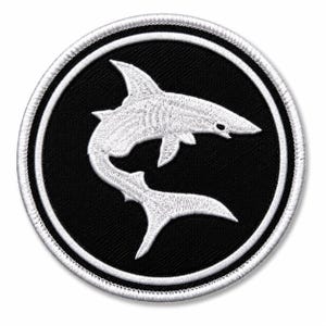 May include: A circular embroidered patch featuring a white shark silhouette centered on a solid black background. The design is enclosed within a thick white border, with a stitched edge finish that creates a clean, defined circular shape.