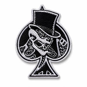 May include: A black and white embroidered patch shaped like a spade playing card suit. The design features a skull wearing a top hat, with intricate patterned details filling the interior space of the spade shape and a white stitched border.