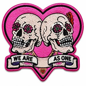 Peut inclure: Un écusson brodé en forme de cœur rose représentant deux crânes blancs avec des orbites florales roses et vertes. Une bannière blanche en bas porte le texte noir WE ARE AS ONE, avec un petit cœur rouge centré sous la bannière.