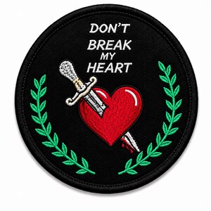 May include: A circular black embroidered patch featuring the white text DON'T BREAK MY HEART at the top. Below the text is a red heart pierced by a silver dagger with a white handle. The design is framed by two green laurel leaf branches.