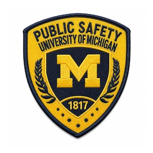 Puede incluir: Un parche bordado en forma de escudo con el texto PUBLIC SAFETY UNIVERSITY OF MICHIGAN en azul oscuro sobre un fondo amarillo. El centro muestra una letra M grande de color amarillo, ramas de laurel y el año 1817 sobre un campo azul oscuro.