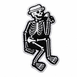Puede incluir: Un parche bordado que muestra un esqueleto en blanco y negro con un sombrero fedora. El esqueleto aparece en posición sentada, sosteniendo una pequeña taza o vaso cerca de la boca. El diseño tiene un borde blanco grueso y costuras de alto contraste.