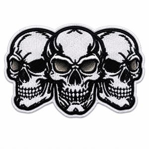 Puede incluir: Un parche bordado que presenta tres calaveras blancas con detalles y contornos negros. Las calaveras están dispuestas una al lado de la otra, con la calavera central mirando hacia adelante y las dos laterales inclinadas hacia afuera, todo con un borde blanco.