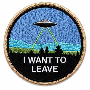 May include: A circular embroidered patch with a tan border featuring a blue sky, green mountains, and a black foreground. A silver UFO hovers above, emitting two green laser beams downward. The text I WANT TO LEAVE is stitched in white.