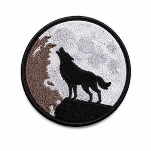 May include: A circular embroidered patch featuring a black wolf silhouette howling at a large white moon. The design includes a brown rocky cliff edge in the foreground and a textured white background, all enclosed within a thick black stitched border.