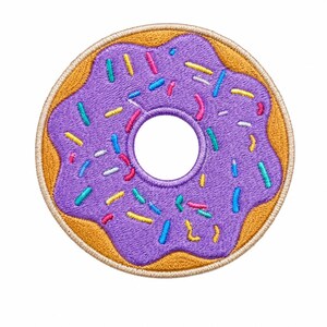 May include: A circular embroidered patch shaped like a donut with golden-brown dough and purple icing. The donut is decorated with small, multi-colored sprinkles in shades of blue, yellow, pink, and white, featuring a stitched border edge.