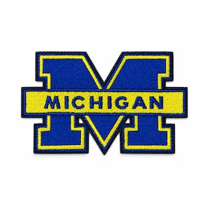 Pode incluir: Um emblema com a letra M em bloco, com fundo azul e borda amarela. Uma barra horizontal amarela atravessa o centro da letra M, contendo a palavra MICHIGAN em letras maiúsculas azuis. O emblema possui uma textura bordada.