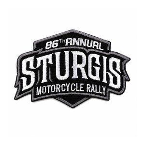 Puede incluir: Un parche bordado en blanco y negro con forma de escudo. El texto dice 86TH ANNUAL STURGIS MOTORCYCLE RALLY en letras mayúsculas blancas y negritas. El parche presenta un borde negro grueso con un contorno gris fino y costuras blancas.