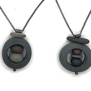 May include: Two necklaces with a dark chain and a pendant featuring a gray circular frame with a stack of small stones in shades of gray and brown. A small stone sits above the frame. The necklaces are displayed against a white background.