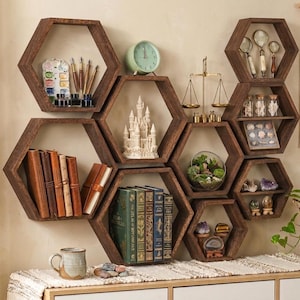 May include: A set of dark brown wooden hexagonal wall shelves arranged in a cluster. The shelves hold various items including books, a white castle figurine, a brass scale, magnifying glasses, glass jars, crystals, a watercolor set, and a green plant.