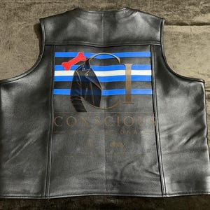 May include: A black leather vest featuring a back panel with horizontal blue and white stripes, a red bone graphic, and a black jacket silhouette. The text CONSCIOUS is printed in gold lettering across the center of the back panel design.