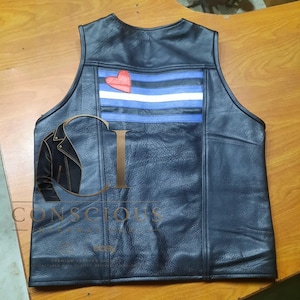 May include: A black leather vest featuring a horizontal striped panel across the upper back in shades of blue and white, accented with a single red heart patch. The vest has a sleeveless design with visible stitching and a textured leather finish.