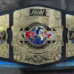 May include: A black leather championship belt featuring a large gold-colored center plate with an engraved world map, international flags, and the text "AEW WRESTLING" and "INTERNATIONAL CHAMPION". Two smaller gold side plates also display the AEW logo.