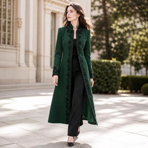 Puede incluir: Un abrigo largo de color verde bosque con cuello alto y un intrincado bordado floral negro a lo largo de la abertura frontal y los puños. El abrigo se lleva sobre una blusa negra sencilla y pantalones negros, creando un aspecto estructurado.