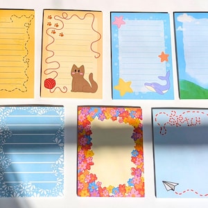 Pode incluir: Sete folhas de papel de carta retangulares com vários designs: uma folha amarela com uma abelha, uma folha bege com um gato e lã, uma folha azul com estrelas-do-mar e uma baleia, uma folha com céu e colinas, uma folha azul floral, uma folha com borda de flores e uma folha com um avião de papel.