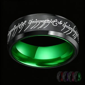 Puede incluir: Un anillo de banda negro con un acabado exterior cepillado y letras grabadas en escritura élfica blanca que rodean la superficie exterior. El interior del anillo tiene un acabado verde vibrante, liso y pulido, creando un contraste llamativo.