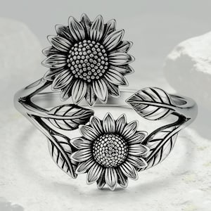 Puede incluir: Un anillo de metal en tono plateado con dos motivos de girasoles y varias hojas. El diseño incluye un girasol grande en la parte superior y uno más pequeño debajo, conectados por una banda curva con texturas detalladas en pétalos y hojas.