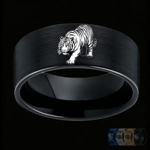 May include: A black ring with a brushed finish featuring a white engraved tiger graphic on the outer band. The ring has a smooth, polished inner surface. Smaller versions of the ring in blue, silver, gold, and black are visible in the corner.
