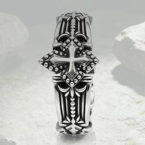 Puede incluir: Un anillo de metal plateado con un prominente motivo de cruz central adornado con pequeñas esferas en relieve. La banda muestra intrincados detalles de ranuras verticales y patrones florales simétricos, con un acabado metálico pulido.