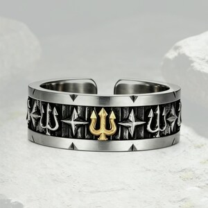 Pode incluir: Um anel aberto de tom prateado com uma banda central escura e texturizada decorada com motivos alternados de tridentes e estrelas prateadas, com um único emblema de tridente dourado centrado na parte frontal do anel.