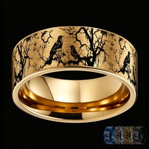 May include: A gold-toned band ring featuring a black laser-engraved design of crows perched on bare, spindly tree branches with small rose blossoms. The exterior surface has a brushed texture, while the interior is smooth and polished gold.
