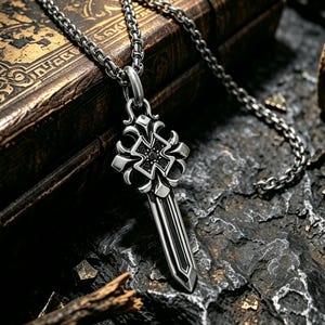 May include: A silver-toned metal pendant shaped like a sword with an ornate cross guard featuring small black stones. The pendant hangs from a matching silver-toned chain link necklace, showcasing a polished finish with dark recessed detailing.