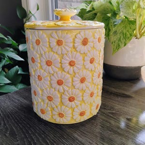 Peut inclure: Un pot cylindrique en céramique de couleur jaune vif, orné d'un motif en relief représentant des marguerites blanches aux cœurs orange texturés. Le pot est surmonté d'un couvercle assorti amovible doté d'un bouton de préhension central jaune.