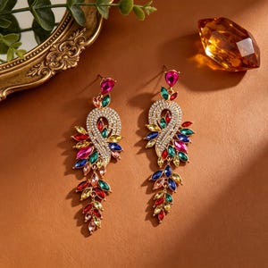 May include: A pair of long, dangling earrings featuring a gold-tone metal frame encrusted with clear rhinestones. The earrings are adorned with multi-colored, marquise-cut crystals in shades of pink, red, green, blue, and yellow in a leaf pattern.