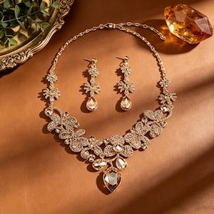 Könnte beinhalten: Ein goldfarbenes Schmuckset bestehend aus einer Halskette und passenden Ohrhängern. Die Halskette ist mit floralen Motiven verziert, die mit klaren Kristallen und bernsteinfarbenen tropfenförmigen Steinen besetzt sind.