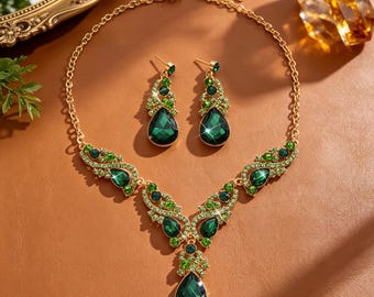 May include: A gold-tone necklace and matching drop earring set featuring dark green teardrop-shaped crystals and smaller light green accent stones. The necklace has a central pendant and symmetrical side motifs, all set in a polished gold-coloured metal.