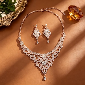May include: A rose gold-toned jewellery set consisting of a necklace and a pair of matching drop earrings. The necklace features an intricate filigree design encrusted with clear crystals, culminating in a central teardrop-shaped pendant element.