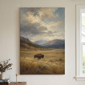 May include: A vertical canvas wall art print featuring an oil painting of a lone brown bison standing in a vast, golden-yellow grassy prairie. In the background, rolling mountains rise under a dramatic, cloudy sky with shades of blue and white.