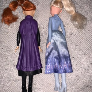 Puede incluir: Dos mu&ntilde;ecas vistas desde atr&aacute;s. La mu&ntilde;eca de la izquierda tiene el pelo rojo en una coleta, una capa morada, mangas negras y botas negras. La mu&ntilde;eca de la derecha tiene el pelo rubio en una coleta, una blusa azul claro y una falda azul estampada.