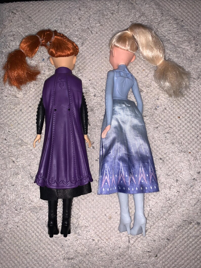 Puede incluir: Dos mu&ntilde;ecas vistas desde atr&aacute;s. La mu&ntilde;eca de la izquierda tiene el pelo rojo en una coleta, una capa morada, mangas negras y botas negras. La mu&ntilde;eca de la derecha tiene el pelo rubio en una coleta, una blusa azul claro y una falda azul estampada.