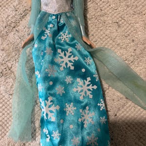 May include: A doll wearing a floor-length turquoise satin skirt featuring a white snowflake and dot pattern. The bodice is white with a subtle glitter finish, complemented by sheer, light blue long sleeves and matching sheer fabric panels.