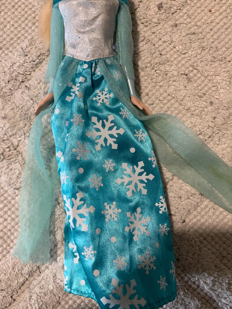 May include: A doll wearing a floor-length turquoise satin skirt featuring a white snowflake and dot pattern. The bodice is white with a subtle glitter finish, complemented by sheer, light blue long sleeves and matching sheer fabric panels.