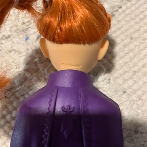 Puede incluir: Una mu&ntilde;eca con cabello naranja brillante peinado en una trenza, con una capa moldeada de color p&uacute;rpura que presenta un sutil dise&ntilde;o floral en relieve en la espalda. La parte posterior del cuello tiene la marca Disney claramente grabada.