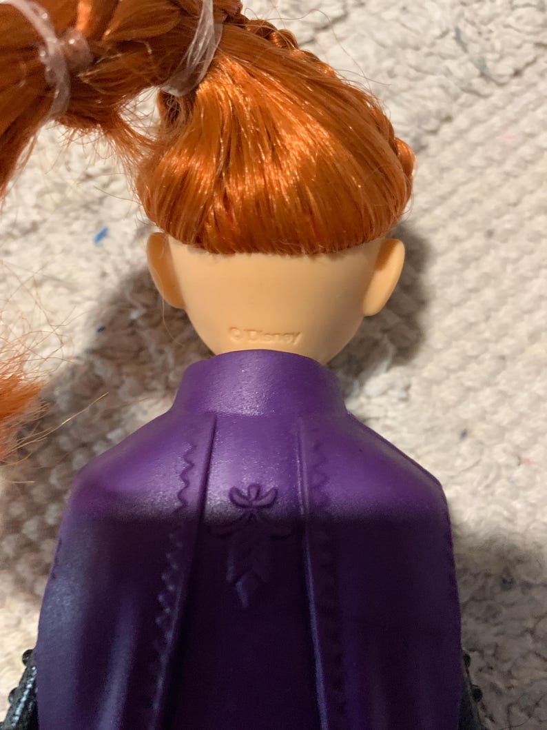 Puede incluir: Una mu&ntilde;eca con cabello naranja brillante peinado en una trenza, con una capa moldeada de color p&uacute;rpura que presenta un sutil dise&ntilde;o floral en relieve en la espalda. La parte posterior del cuello tiene la marca Disney claramente grabada.