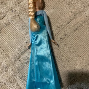 May include: A doll with blonde hair styled in a long braid, wearing a light blue sleeveless gown with a satin-finish skirt and a textured bodice. The doll is viewed from the back, showing the hair braid resting against the upper back area.