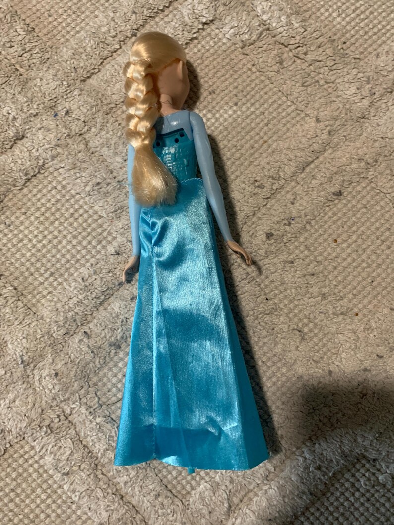 May include: A doll with blonde hair styled in a long braid, wearing a light blue sleeveless gown with a satin-finish skirt and a textured bodice. The doll is viewed from the back, showing the hair braid resting against the upper back area.