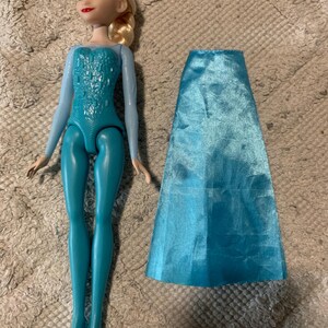 May include: A plastic doll with blonde hair styled in a side braid, blue eyes, and red lips. The doll wears a light blue long-sleeved bodice with a textured pattern and teal leggings. A separate, detachable, shiny light blue satin skirt is included.