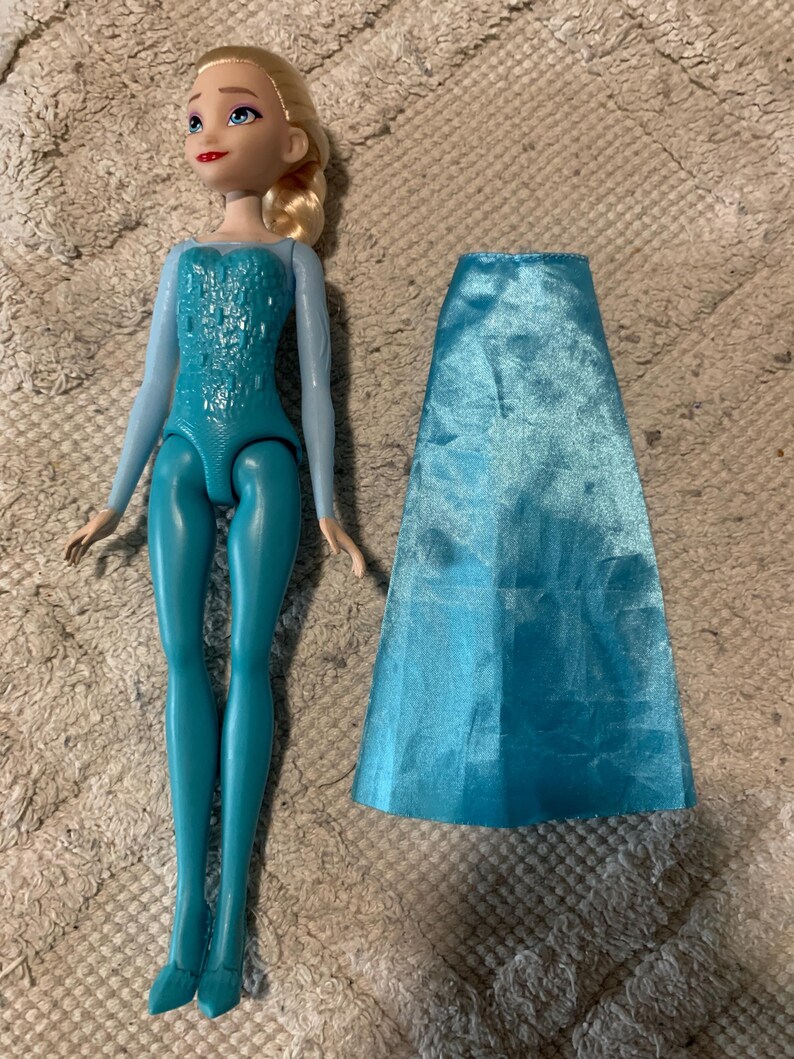 May include: A plastic doll with blonde hair styled in a side braid, blue eyes, and red lips. The doll wears a light blue long-sleeved bodice with a textured pattern and teal leggings. A separate, detachable, shiny light blue satin skirt is included.